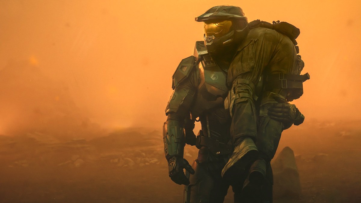 Master Chief  in full armor carries someone over his shoulder against an orange sky in Halo season two