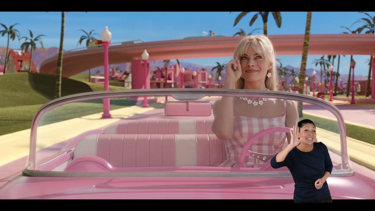 Barbie drives her pink Cadillac and smiles with a woman in black signing the movie in ASL in the bottom righthand corner