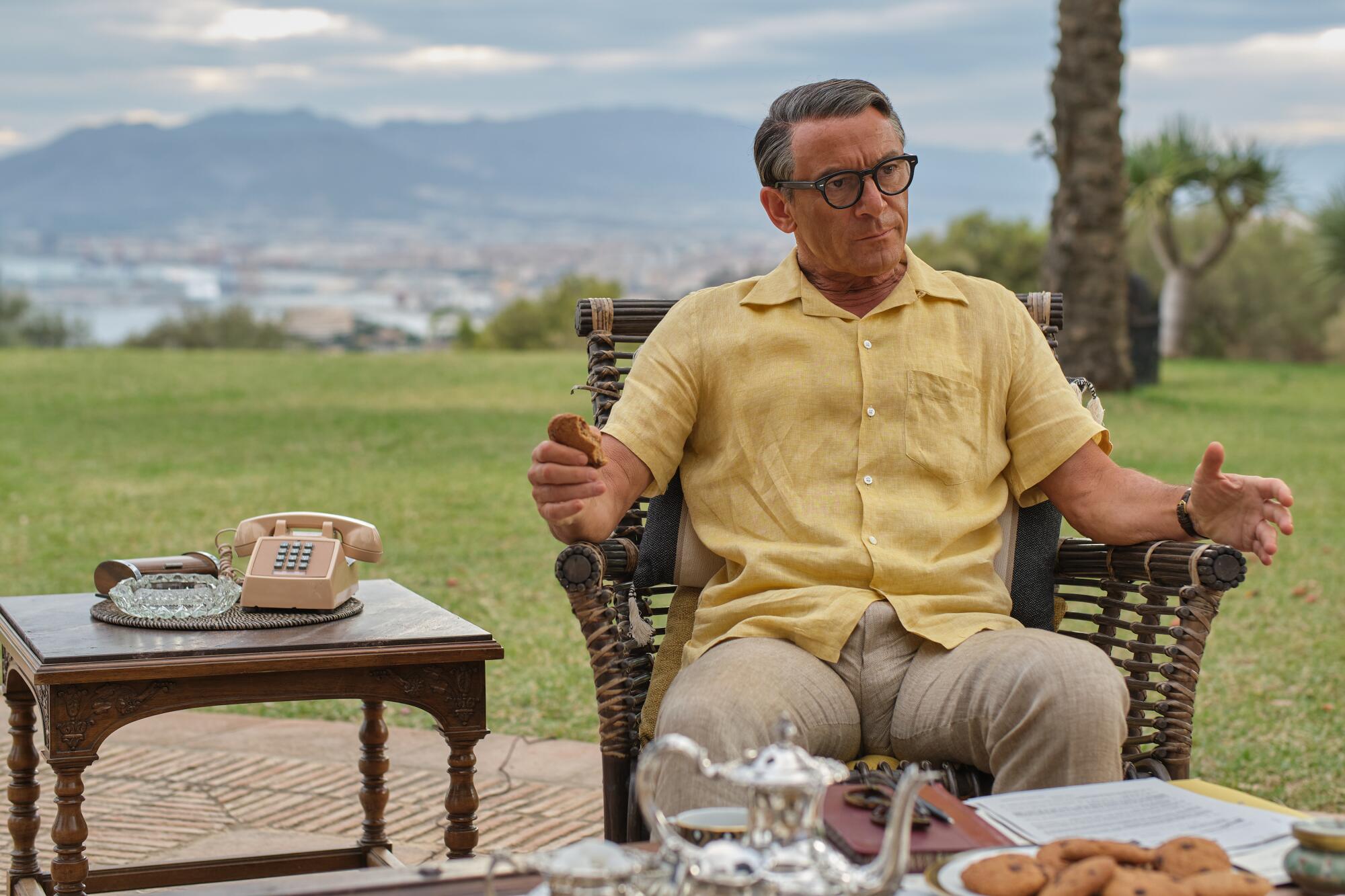 Jason Isaacs as Cary Grant in a yellow shirt and khakis sitting in a chair outside.