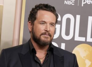 Cole Hauser smiles at a red carpet event, wearing a black suit coat and a black shirt with a gold necklace