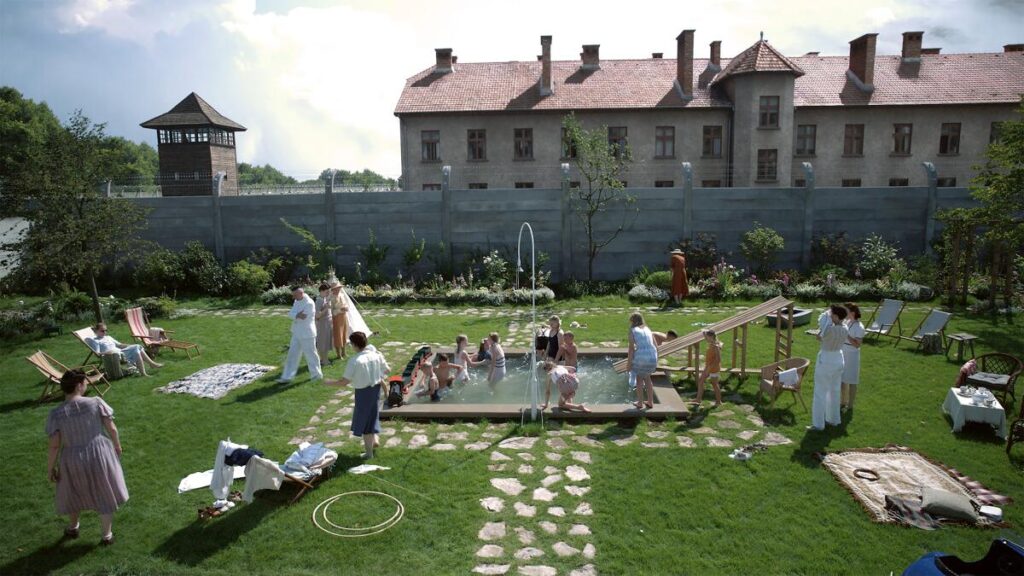 A garden party shows the buildings of Auschwitz just over the backyard wall.