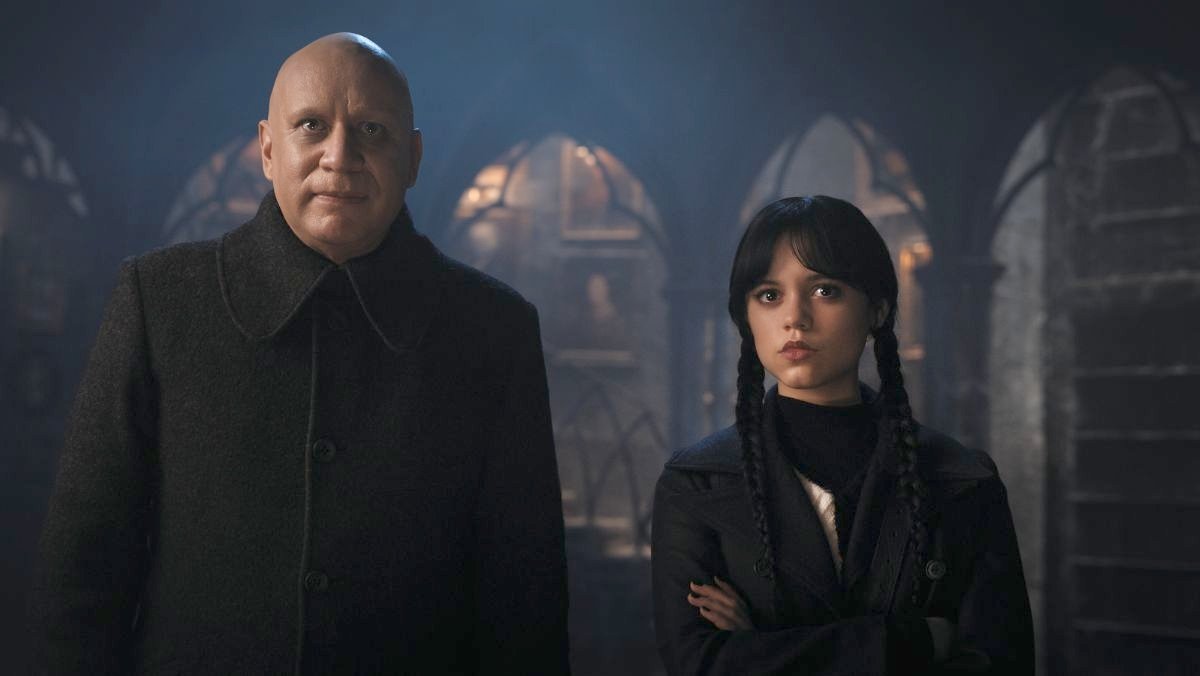 Uncle Fester and Wednesday Addams stand next to each other in a hallway