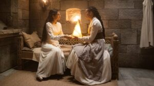 Egwene and Nynaeve holding hands, sitting on a bed inside the White Tower in The Wheel of Time season two