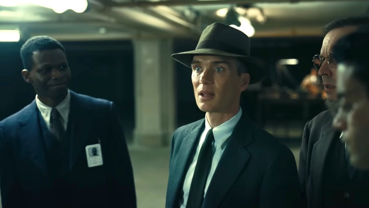 Cillian Murphy in a hat and suit surrounded by other men in Oppenheimer