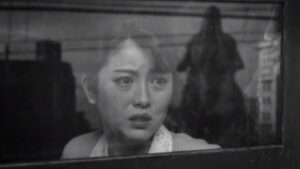 A scared woman looks out the window of a train and sees Godzilla rampaging through the city in Godzilla Minus One in black and white.