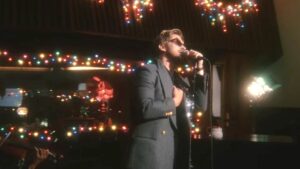Ryan Gosling in sunglasses and a sports coat sings in front of Christmas lights in a studio