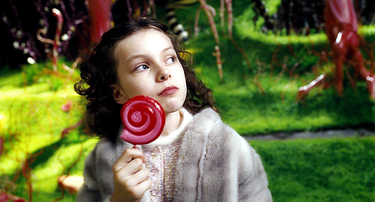 Veruca Salt (Julia Winter), a young girl with curly hair, wearing a fur coat, stands in a garden made of candy, holding a huge, swirly red lollipop, in Tim Burton’s Charlie and the Chocolate Factory