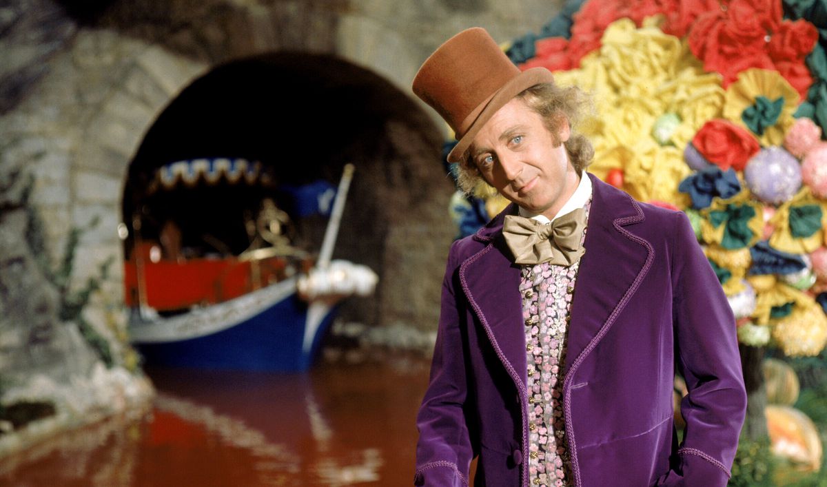 Willy Wonka (Gene Wilder, in purple velvet jacket and tan top hat) stands in front of a chocolate river as a ship appears out of a tunnel behind him in 1971’s Willy Wonka &amp; the Chocolate Factory