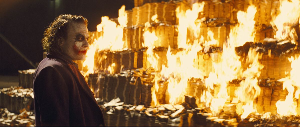 Heath Ledger as Joker stands in front of a vast pile of burning stacks of cash in The Dark Knight