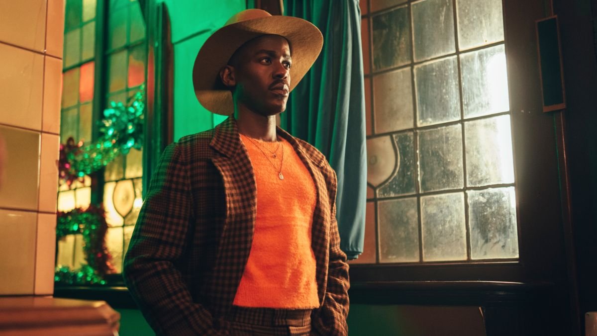 Ncuti Gatwa as the Doctor stands near a glass stained window wearing a checkered brown suit, orange sweater, and a cowboy hat
