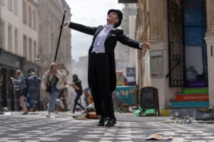 Neil Patrick Harris dances in a tux with coattails, a top hat, and cane as chaos breaks out on the streets of London behind him in the Doctor Who special “The Giggle”