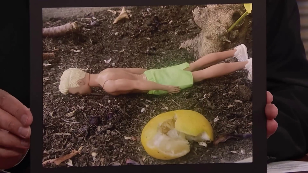 A close-up of Jimmy Fallon’s hands holding up a photo of a Ken doll, face down in the dirt, next to a squished lemon