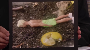 A close-up of Jimmy Fallon’s hands holding up a photo of a Ken doll, face down in the dirt, next to a squished lemon