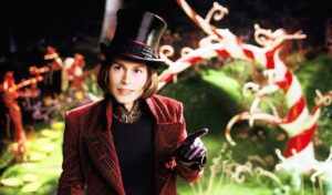 Willy Wonka (Johnny Depp, in red velvet suit jacket and glossy black top hat) stands smiling in front of a garden made of candy in Tim Burton’s 2005 Charlie and the Chocolate Factory