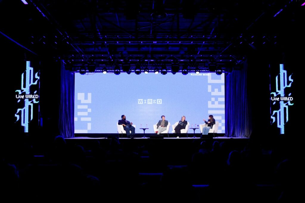 Four people sitting on a stage in a dark room with a large screen behind them showing WIRED logos
