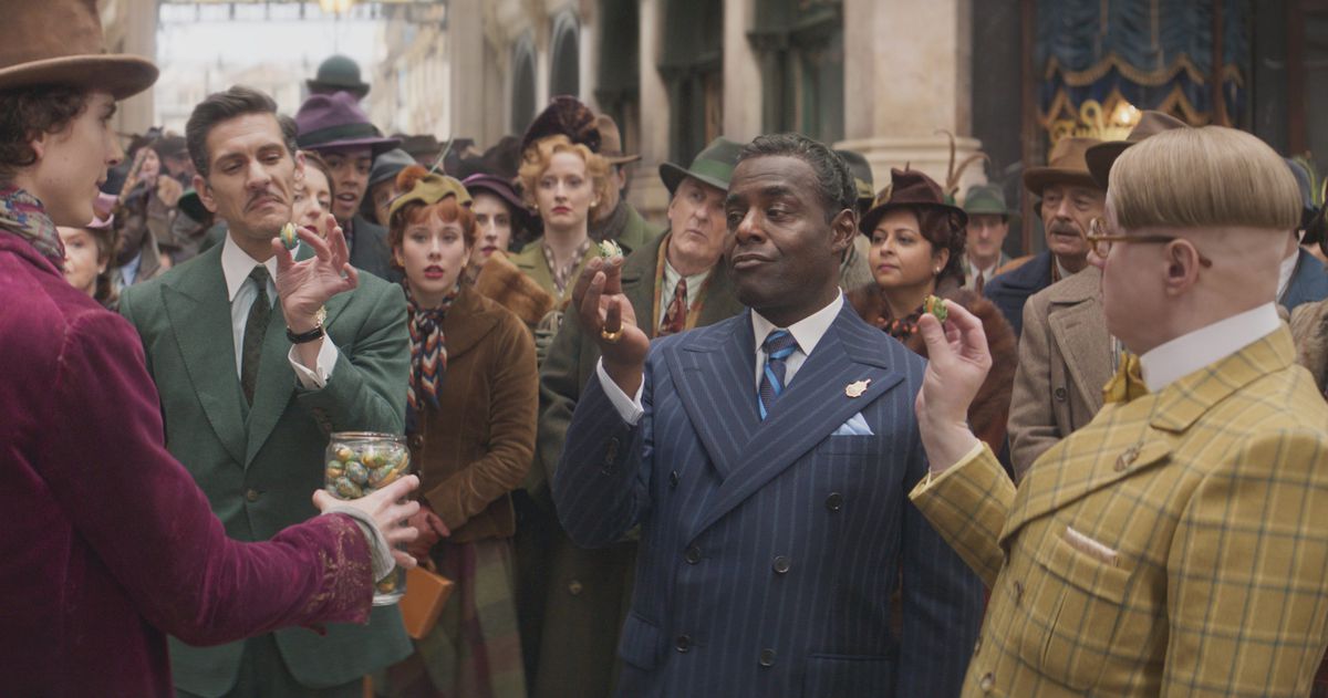 Mathew Baynton in a green suit, Paterson Joseph in a blue suit, and Matt Lucas in a yellow suit talking to Willy Wonka