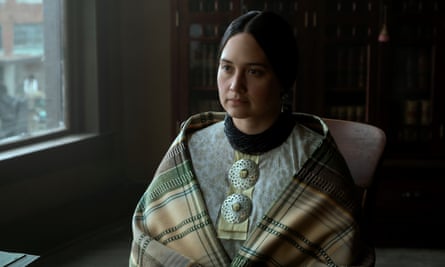 A Native American woman with long black hair parted in the middle sits indoors wrapped in a blanket.