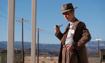 A middle-aged white man wearing a brimmed hat and trench coat, one hand on his hip, holds a pipe in the other as he looks at a desert-ish landscape  beyond telephone poles.