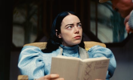 Young white woman with black hair parted in the middle, dressed like Alice in Wonderful in a large-shouldered powder blue dress, holding a book as she sits in a chair.