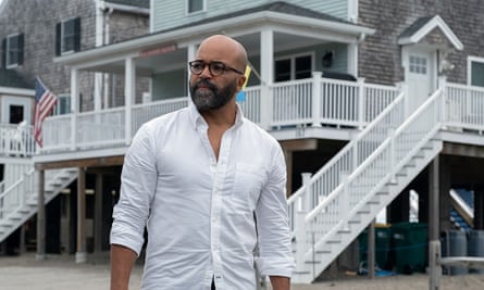Bald Black man with trim beard in tortoiseshell glasses and white button-down Oxford, standing on maybe a beach in front of a large house with a wraparound porch.