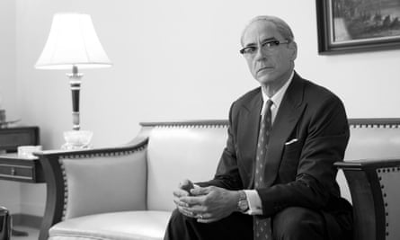 White man with bald head and glasses, wearing suit, sits on nice sofa looking serious.