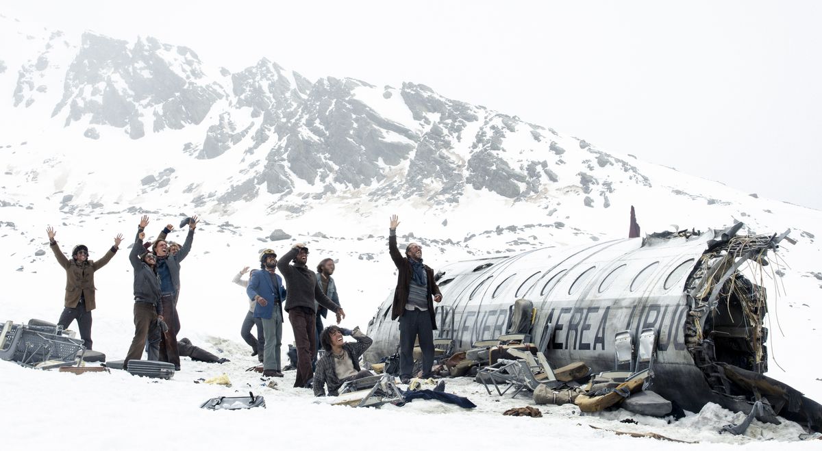 A group of survivors, seen as tiny figures from a long distance away, stand in the snow next to their fragmented plane and wave at something offscreen in Netflix’s Society of the Snow.