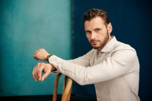 Adan Canto resting his hands on a wooden chair, dressed in an off-white shirt