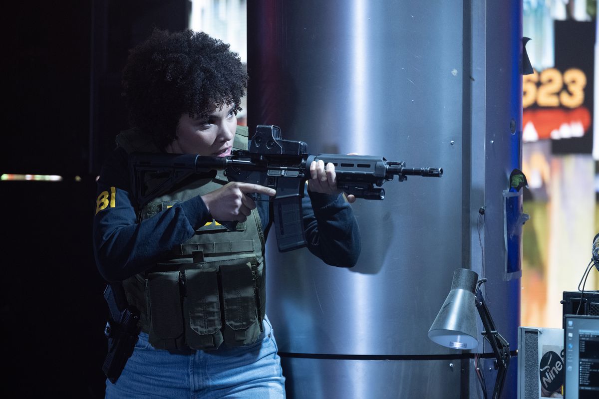 Emmy Raver-Lampman, wearing a bulletproof vest and an FBI uniform, aims an assault rifle in The Beekeeper.