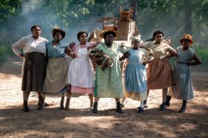 A line of seven women in various muted pastel dresses and aprons in a scene from "The Color Purple."