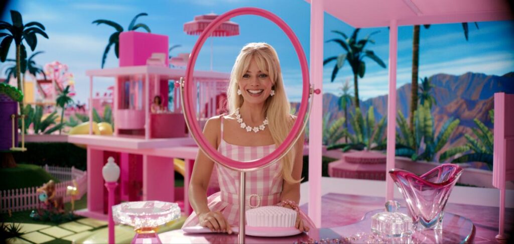 Margot Robbie as Barbie smiles in front of a mirror inside a pink doll house.