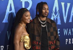 Halle Bailey with braids in a gold top posing next to DDG in a brown-checkered jacket against a blue backdrop