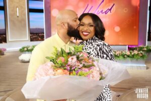 Common in a pastel yellow outfit kissing Jennifer Hudson, wearing glasses and a black-and-white patterned dress, on the cheek