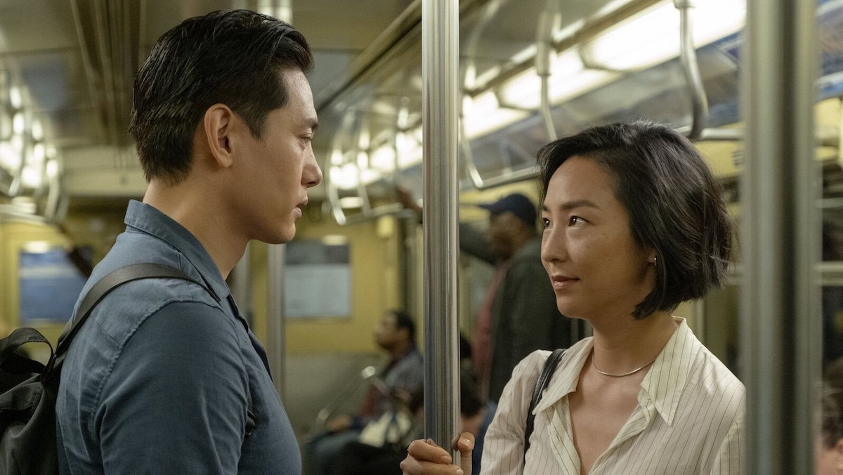 A man in a blue shirt looks at a woman holding the pole on a train in Past Lives