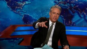 jon stewart at the daily show desk pointing at the camera