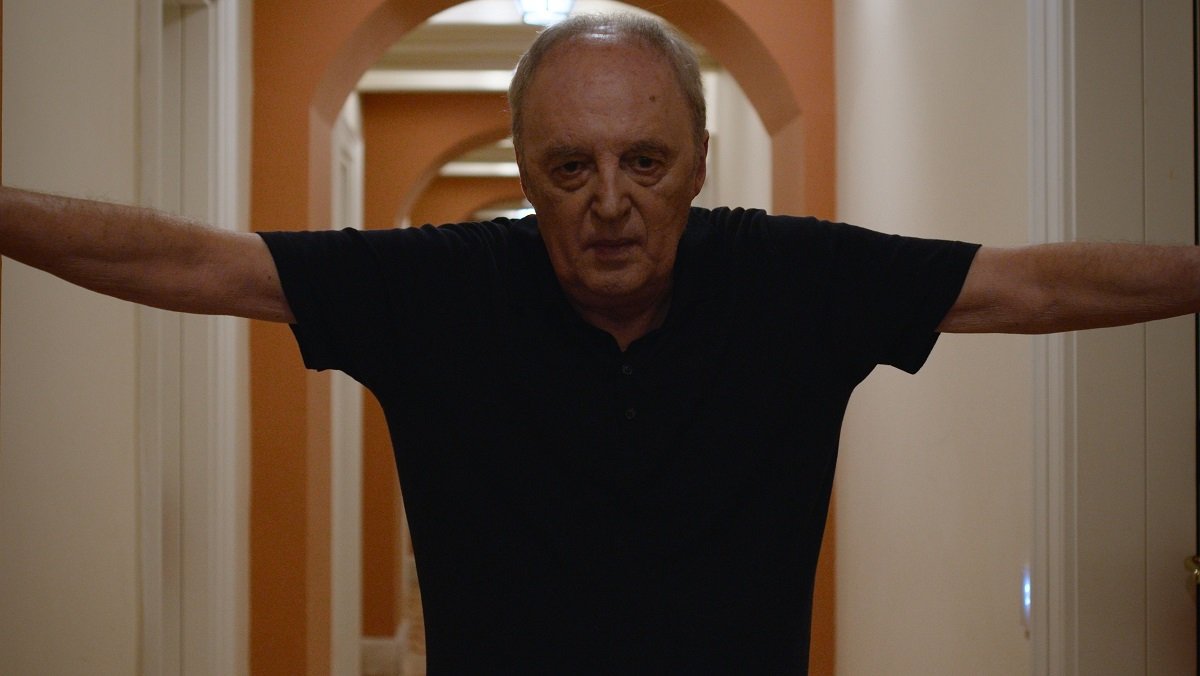Dario Argento stands in a long hallway, his hands outstretched on the walls, in the documentary Dario Argento Panico.