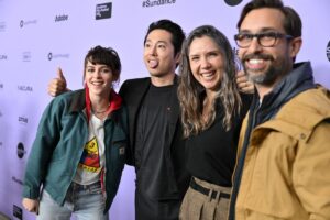 Kristen Stewart, Steven Yeun, Sam Zuchero, and Andy Zuchero pose in front of a Sundance backdrop, with Yeun sticking out his tongue and giving a thumb’s up, at the Sundance premiere of their movie Love Me