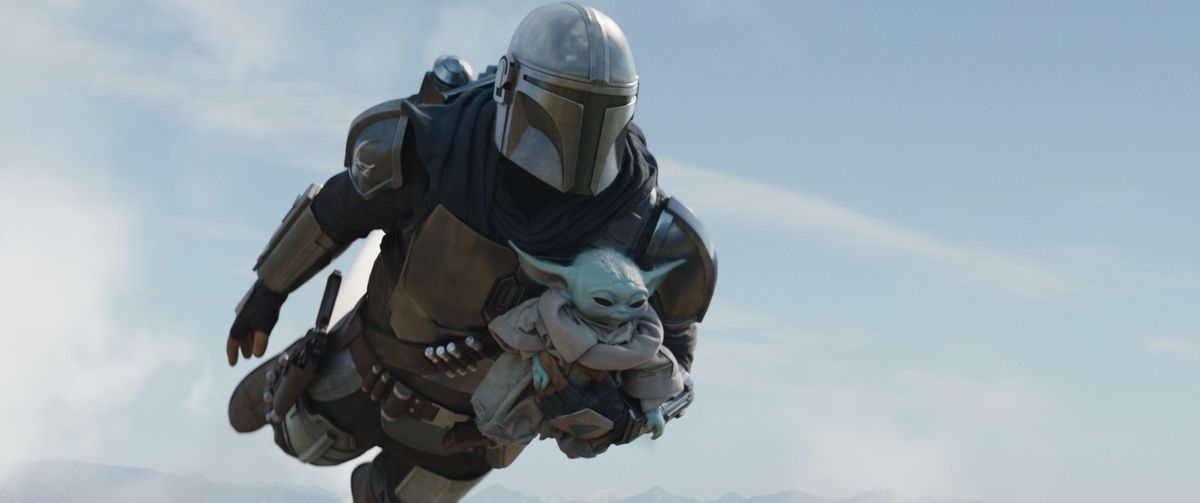 The Mandalorian, Din Djarin, holding Grogu in his arms while flying with a jetpack.