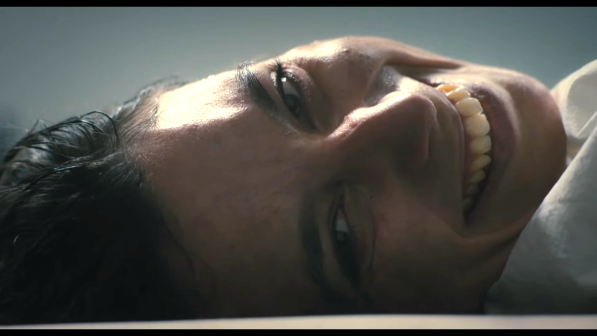 a woman lies on her back and faces the camera with an ominous smile in the first omen trailer
