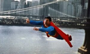 Man wearing blue leotard and red cape flies in front of a bridge and over water.