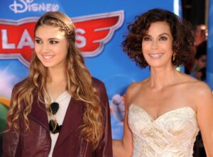 A young woman in a jacket and a braided hairstyle stands next to an older woman with short curly hair in a strapless dress