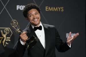 Trevor Noah holds a Primetime Emmy in one hand and gestures with the other while smiling in a tux backstage at an awards show