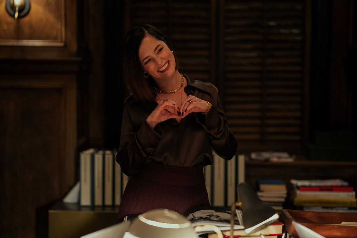 Parker Posey smiles and makes a love heart shape with her hands in a dark study in Mr. &amp; Mrs. Smith