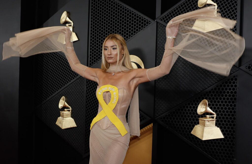 A woman in a beige bustier dress adorned with a yellow ribbon raises her arms on the red carpet