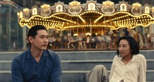 A man and woman sit near a carousel in "Past Lives."