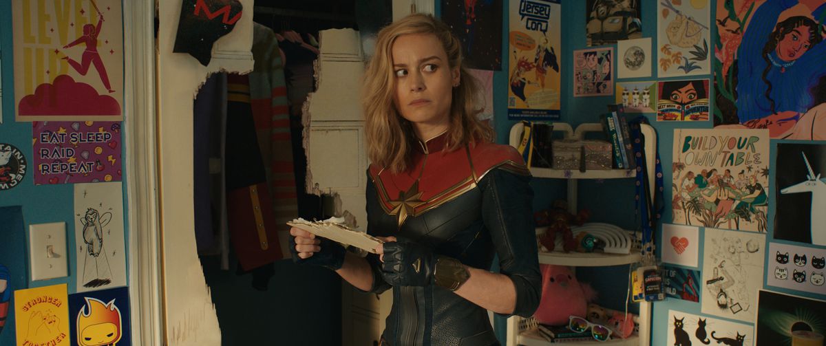 Captain Marvel (Brie Larson) stands in Kamala Khan’s bedroom next to a smashed closet door, looking around with alarm at all the cartoon images of herself, in the Marvel Cinematic Universe movie The Marvels