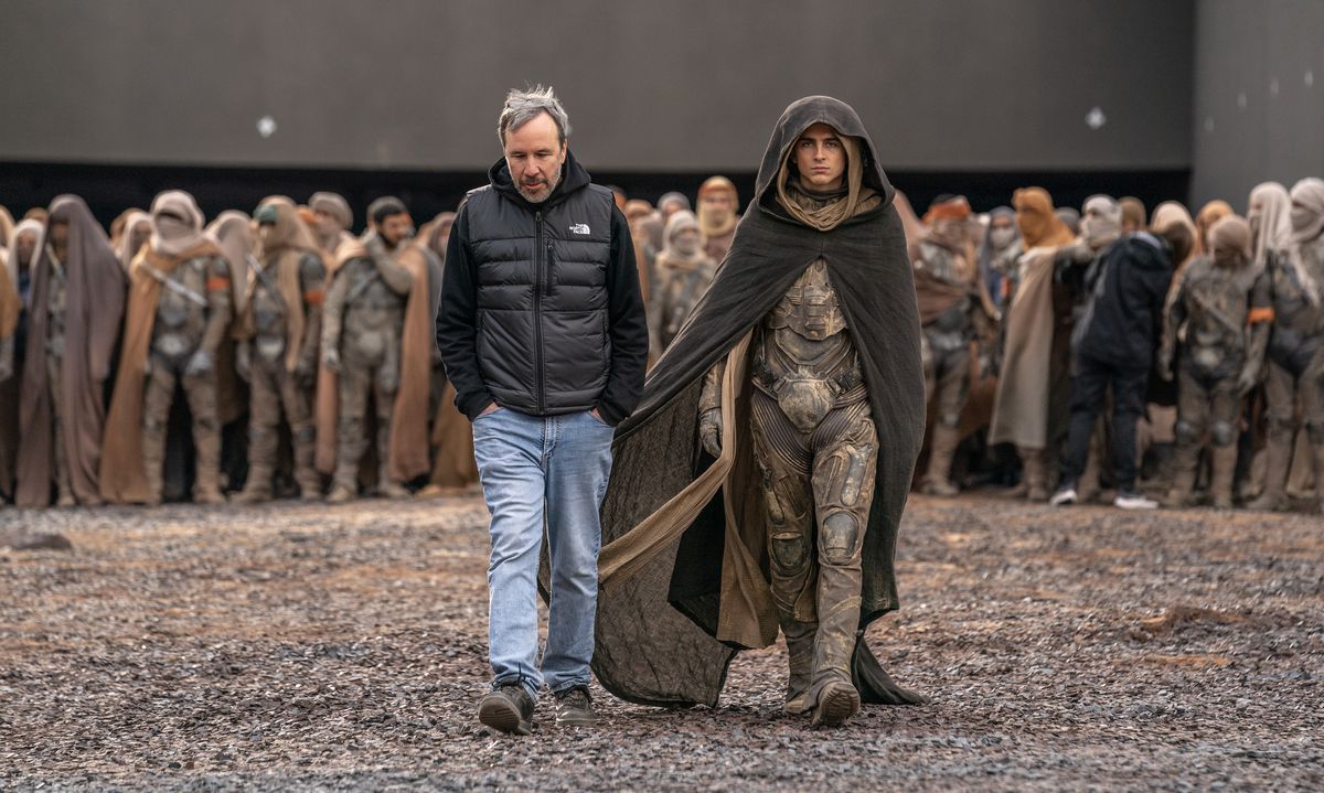 Denis Villeneuve and a costumed Timothée Chalamet walk across a gravel field on the set of Dune Part 2, a large group of extras costumed as Fremen behind them.