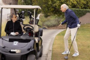 Richard Lewis sits in a golf cart looking at Larry David, who stands on the grass holding a golf club.