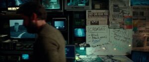 John Krasinski sits out of focus at a computer, the camera showing us a whiteboard with lots of clues to the aliens and newspaper clippings about the invasion, in A Quiet Place