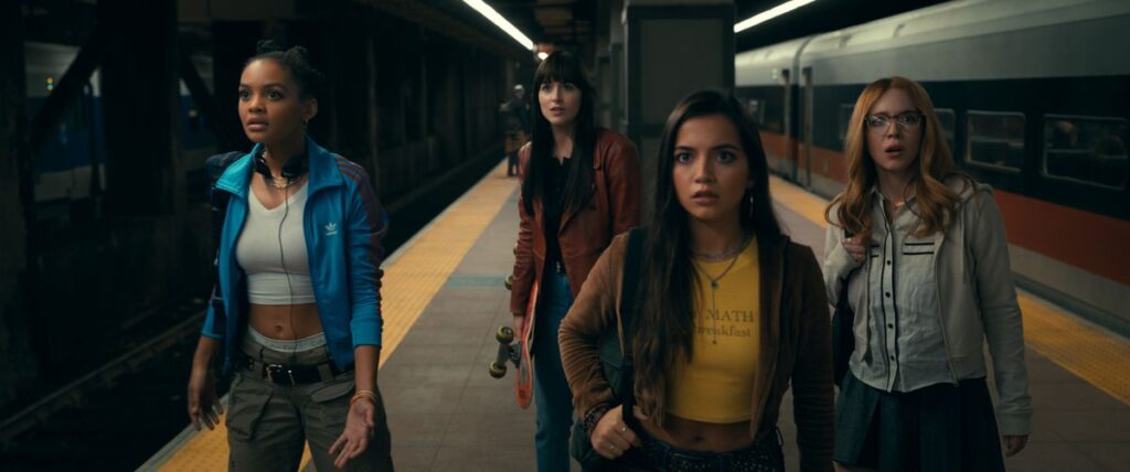 Three young women stand on a subway platform, looking shocked. An adult woman stands slightly behind them, on guard.