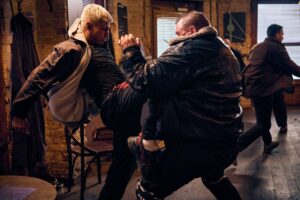 A man with blond hair performs a high-knee strike into another man’s abdomen while another man runs away in the distance.